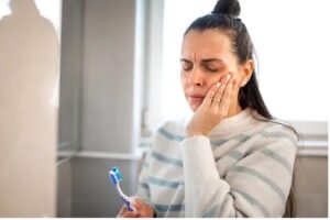 Woman grimacing holding her jaw with a toothbrush in her hand.